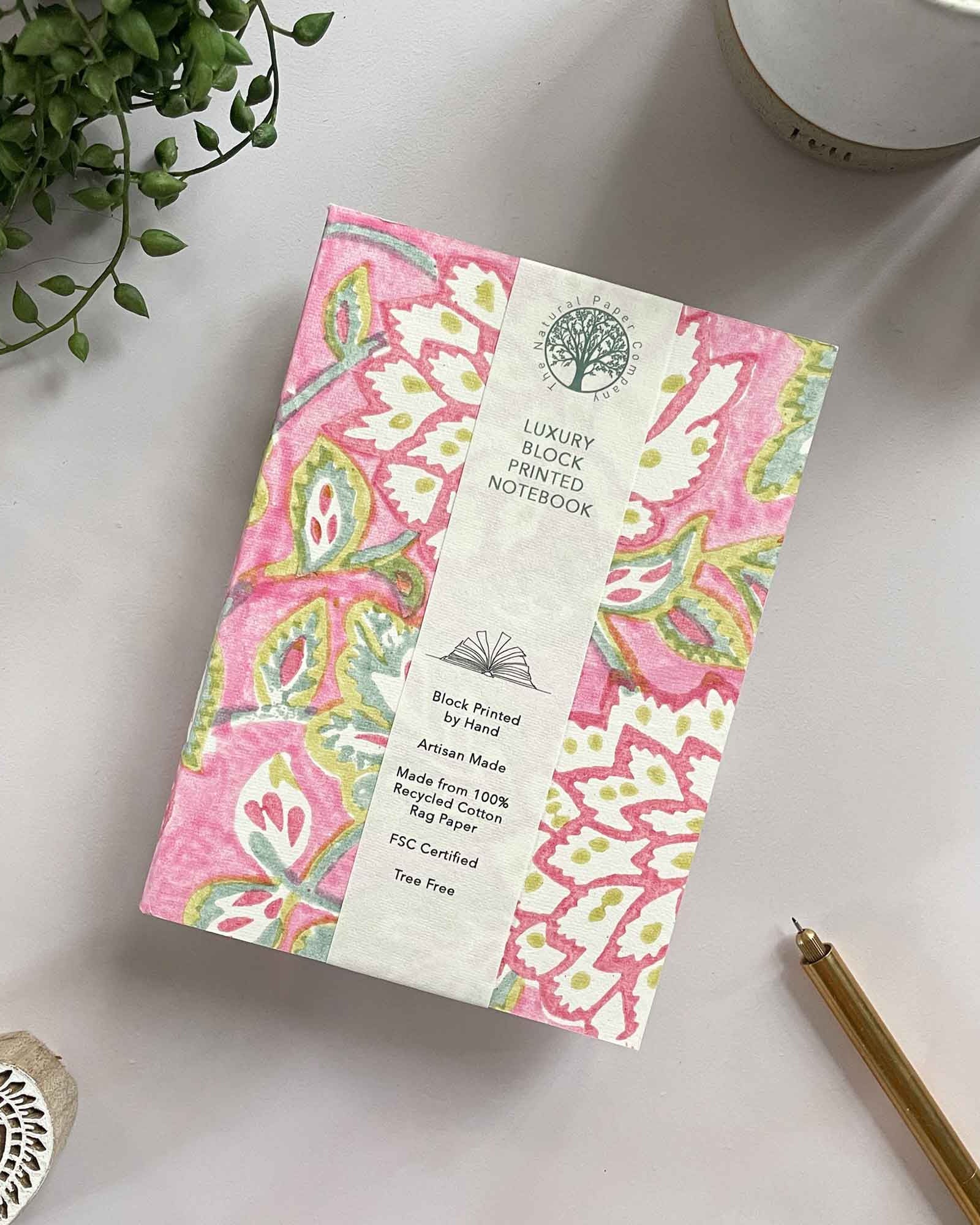 Decorative notebook with pink floral pattern on a white surface with a plant and pen.