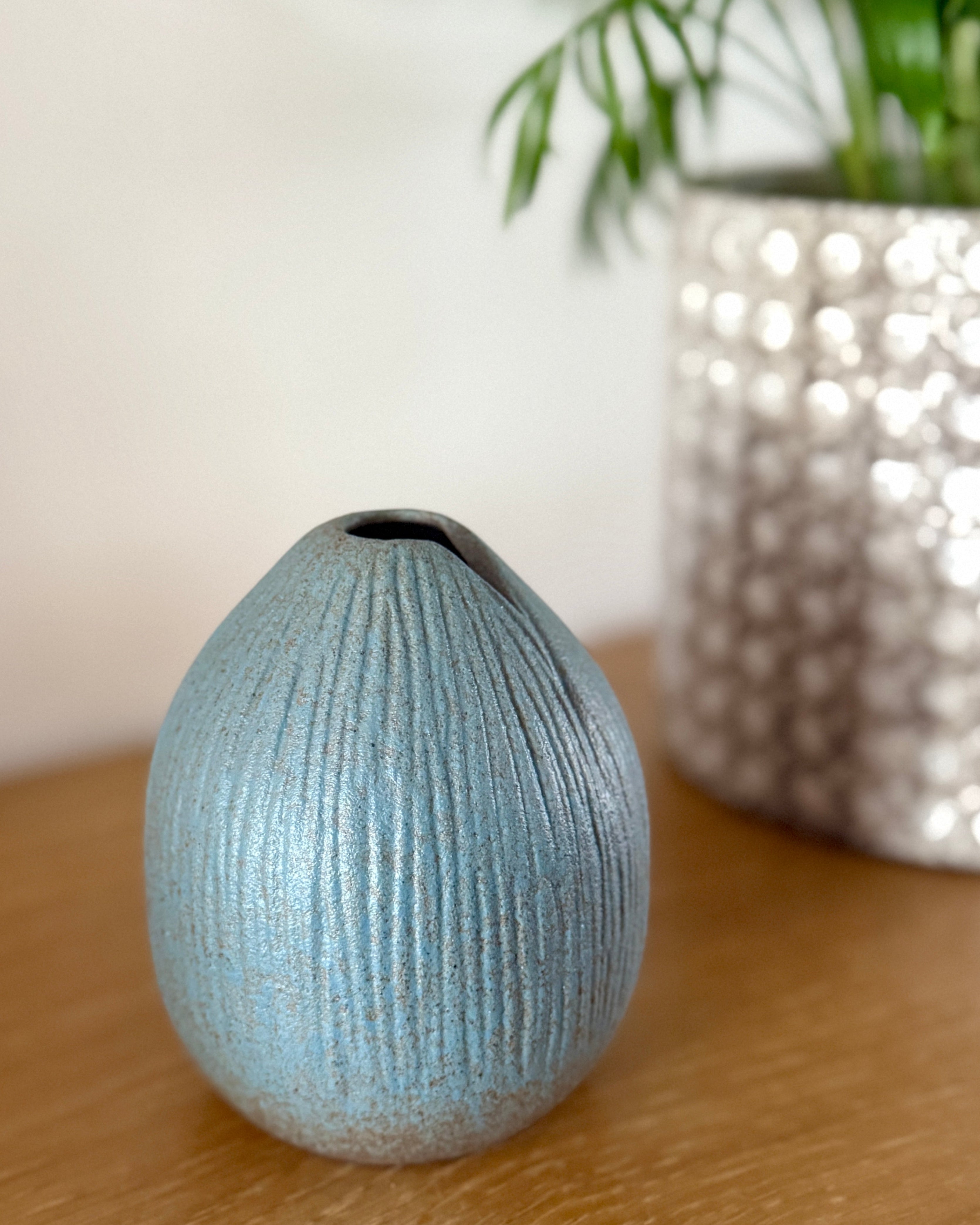 Textured blue vase on a wooden surface with a blurred silver pot and plant in the background.