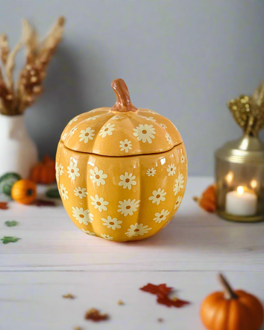 Yellow pumpkin-shaped container with floral design on a wooden surface, next to pink spiral candles.