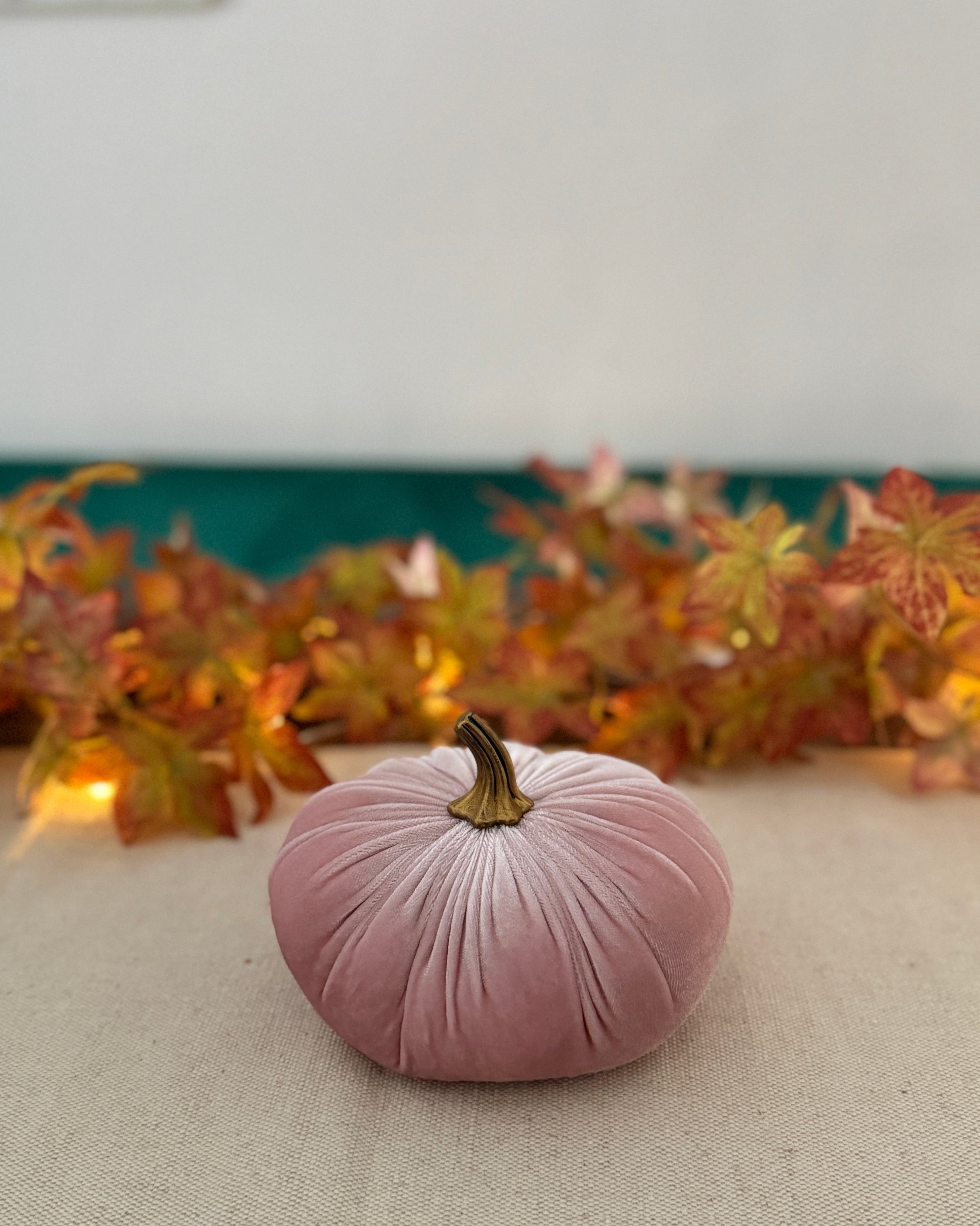 Pink velvet pumpkin on a neutral surface with autumn leaves in the background