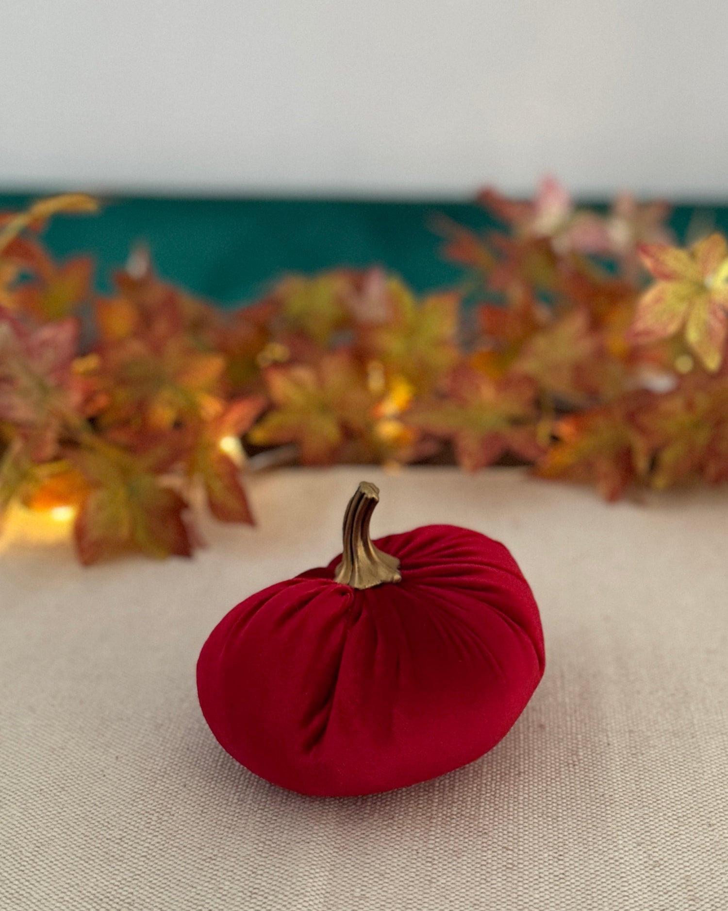 Red decorative pumpkin with a gold stem on a beige surface with autumn leaves in the background.