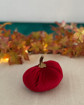 Red decorative pumpkin with a gold stem on a beige surface with autumn leaves in the background.