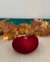 Red decorative pumpkin with a gold stem on a textured surface with autumn leaves in the background.