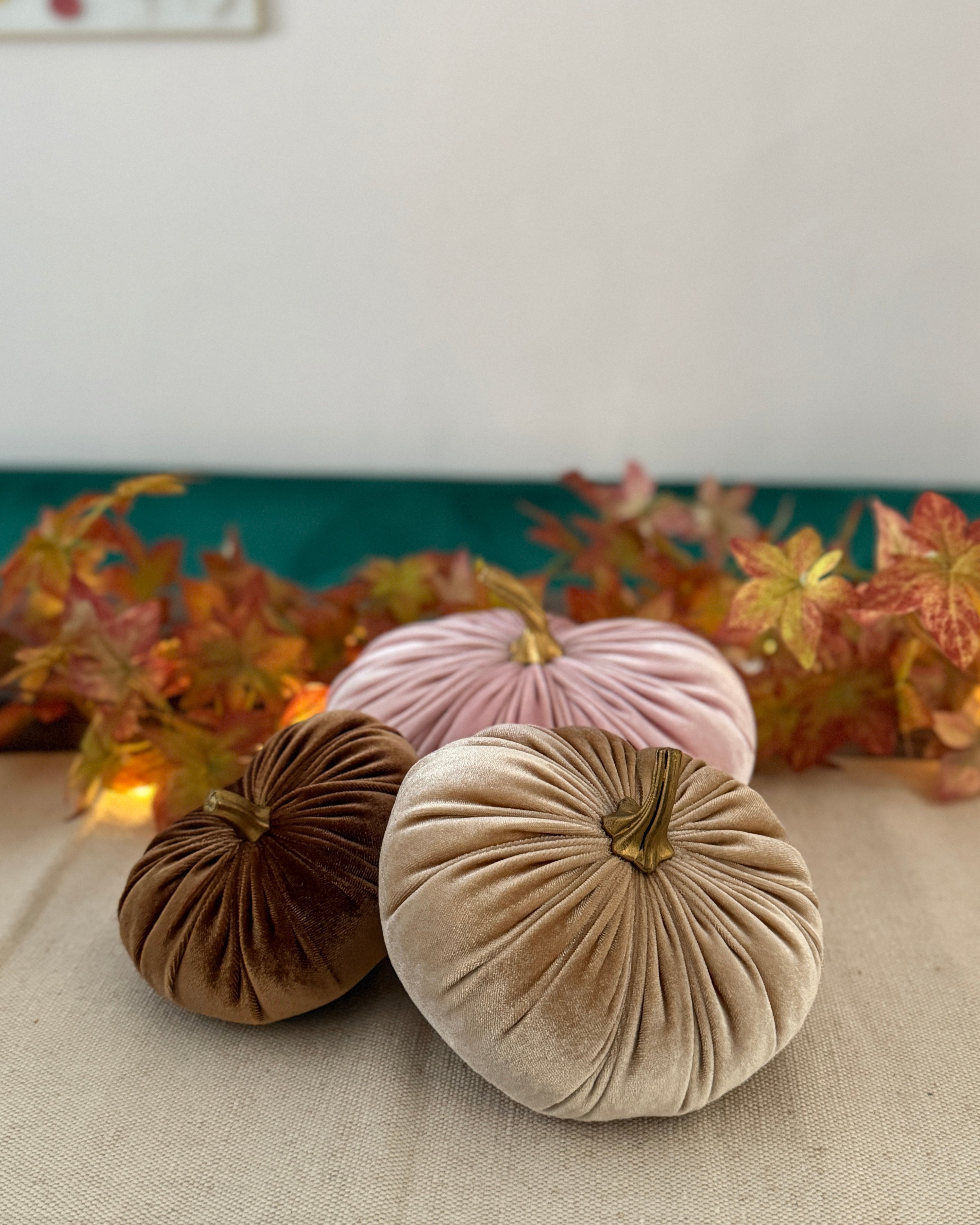 Three velvet pumpkins in brown, pink, and beige on a table with autumn leaves in the background.
