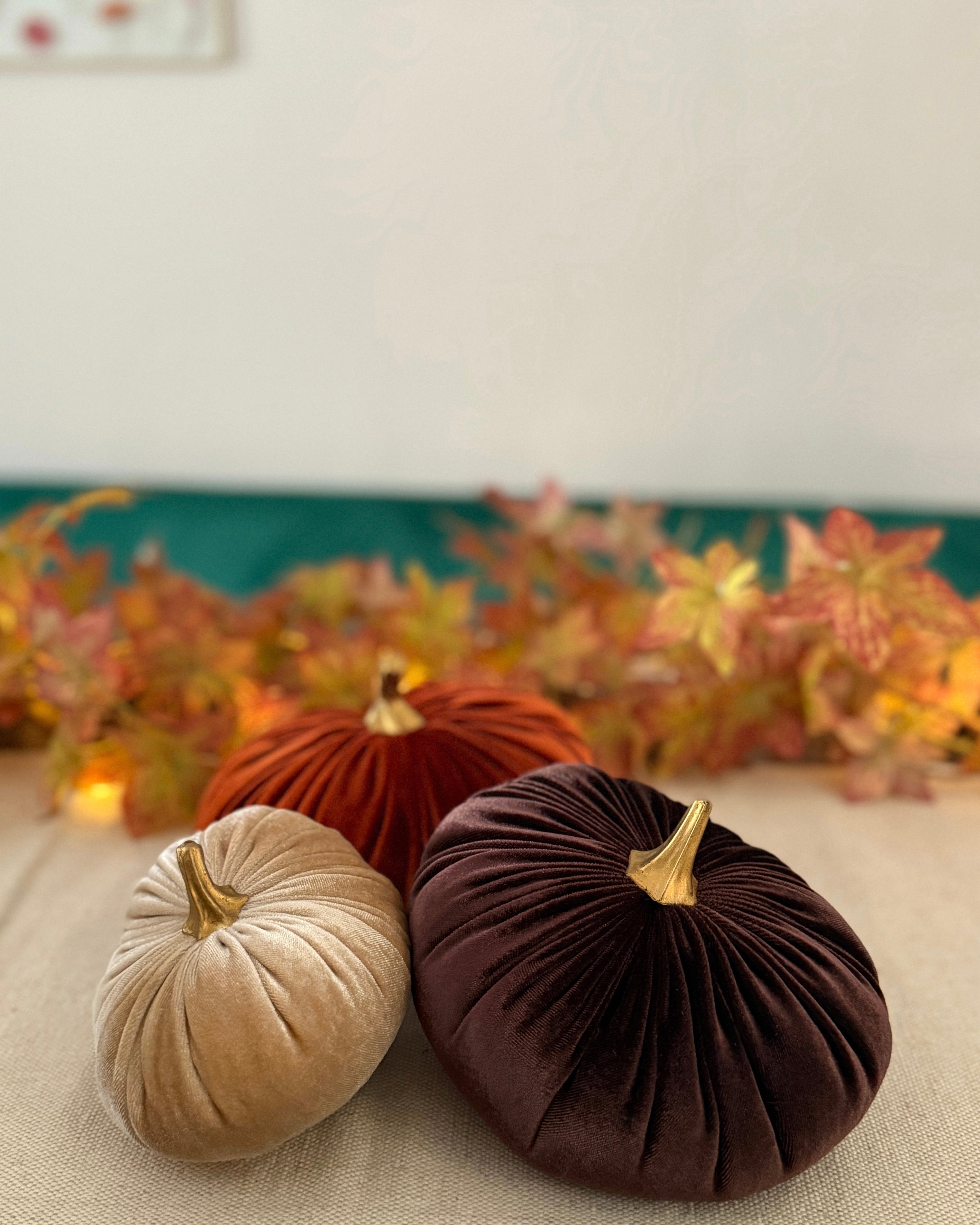 Three velvet pumpkins in beige, brown, and red on a neutral surface with a decorative leaf garland in the background.