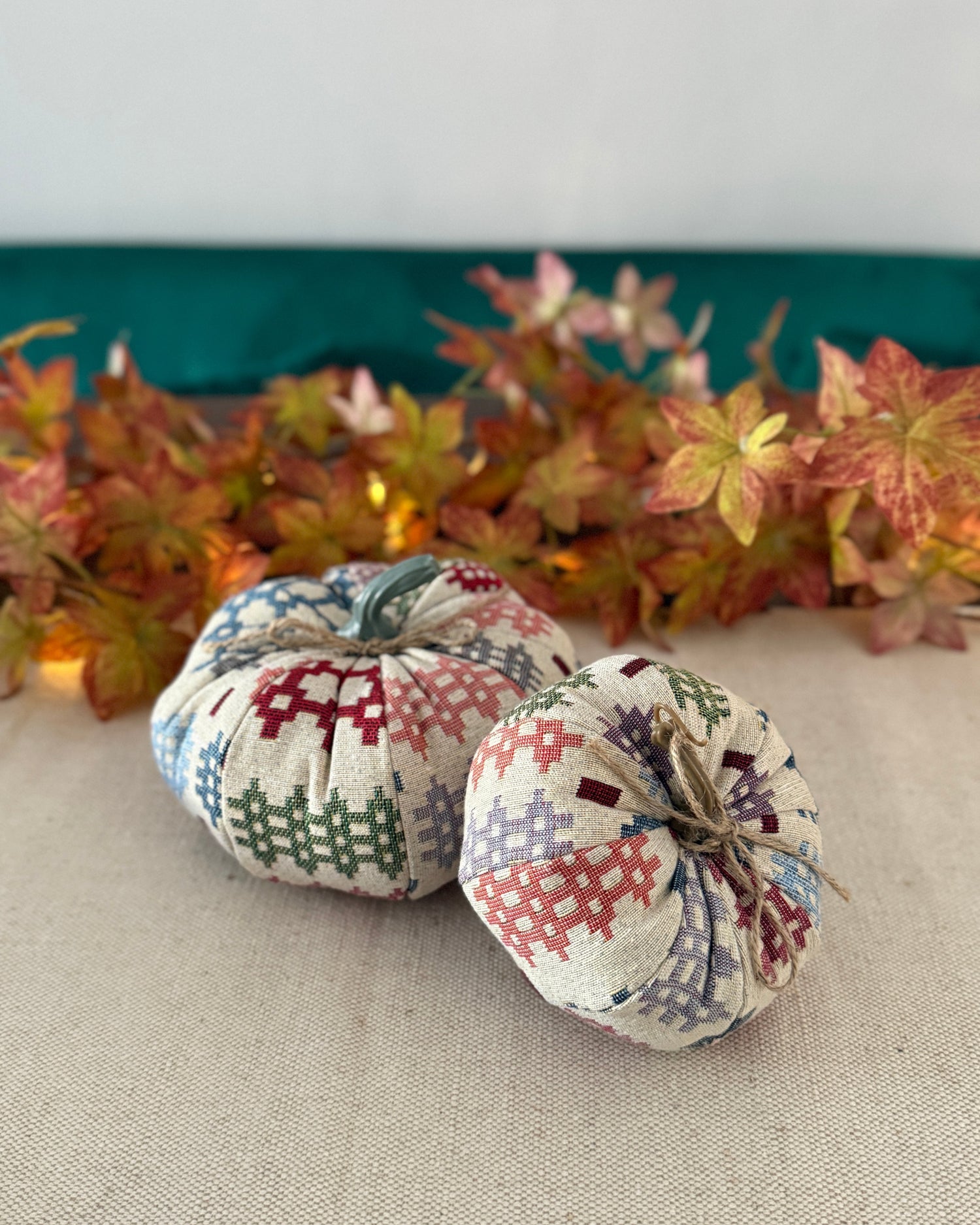 Two patchwork-style fabric pumpkins on a neutral surface with autumn leaves in the background.
