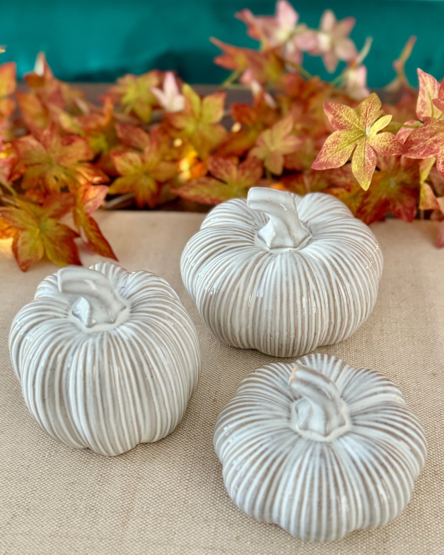 Three white textured pumpkin decorations on a beige surface with autumn leaves in the background.