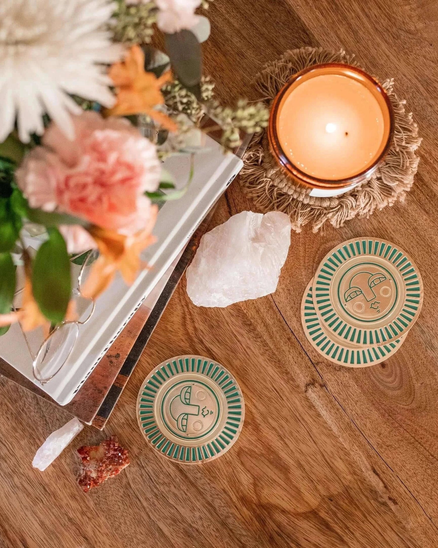 handmade teal coasters with gold-coloured metal and enamel, featuring the Sol face design. Coasters placed on a table setting with flowers, candle and books
