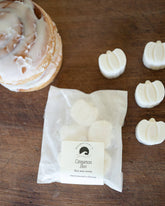 Packaged cinnamon bun-shaped bath bombs on a wooden surface with a cinnamon roll.