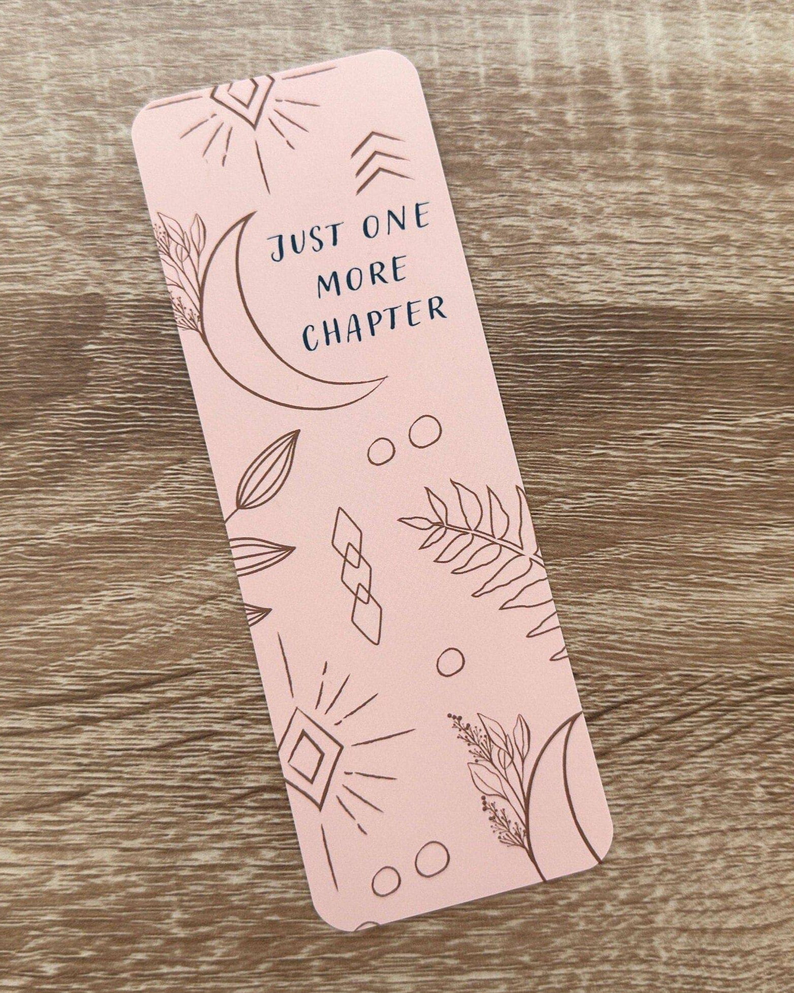 Boho-themed waterproof bookmark with "Just One More Chapter" text, featuring illustrations of moons, leaves, and abstract shapes in pink and brown tones. Made from recycled card with a velvet lamination.