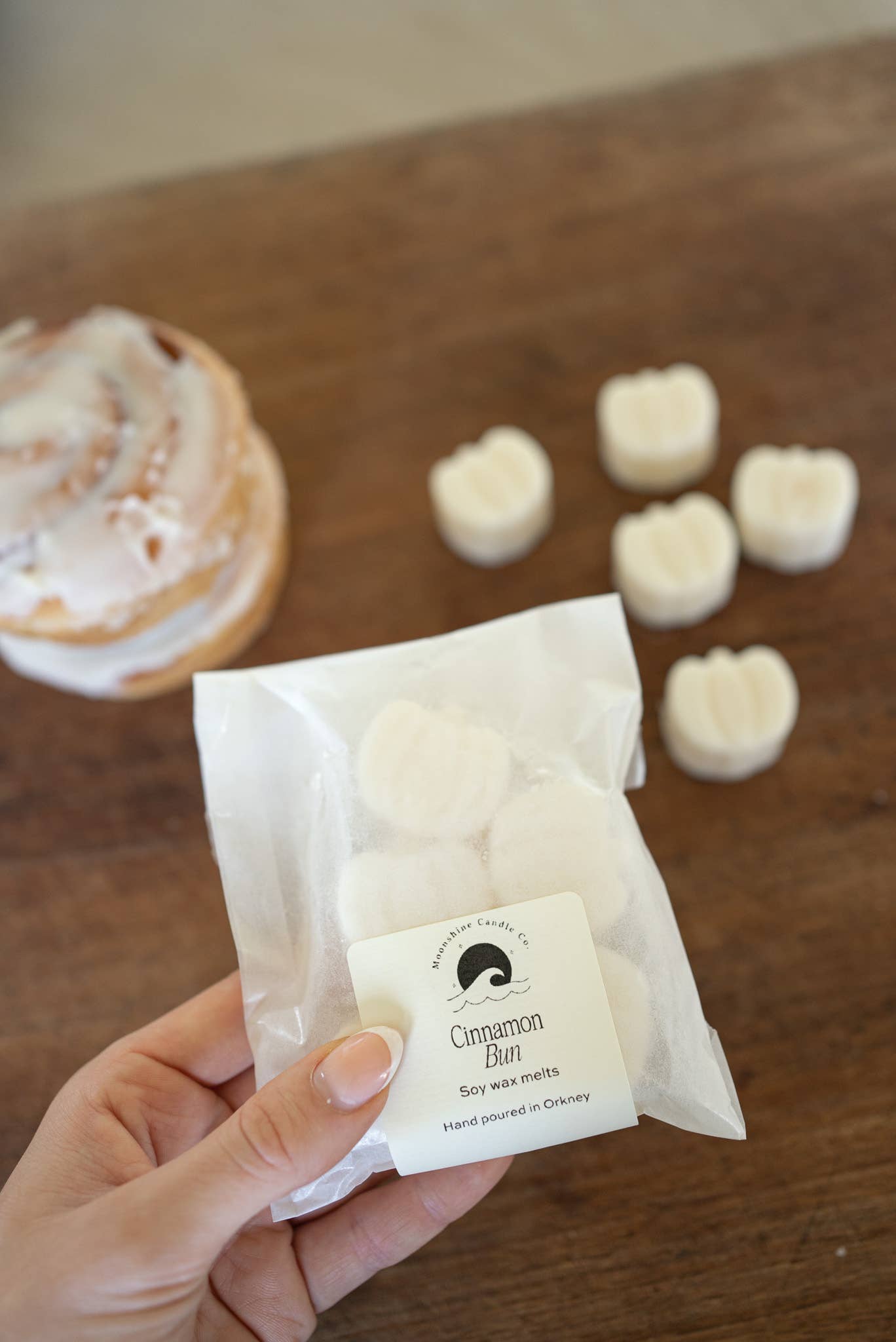 Hand holding a small package of cinnamon bun-flavored candy on a wooden surface with a cinnamon roll in the background.