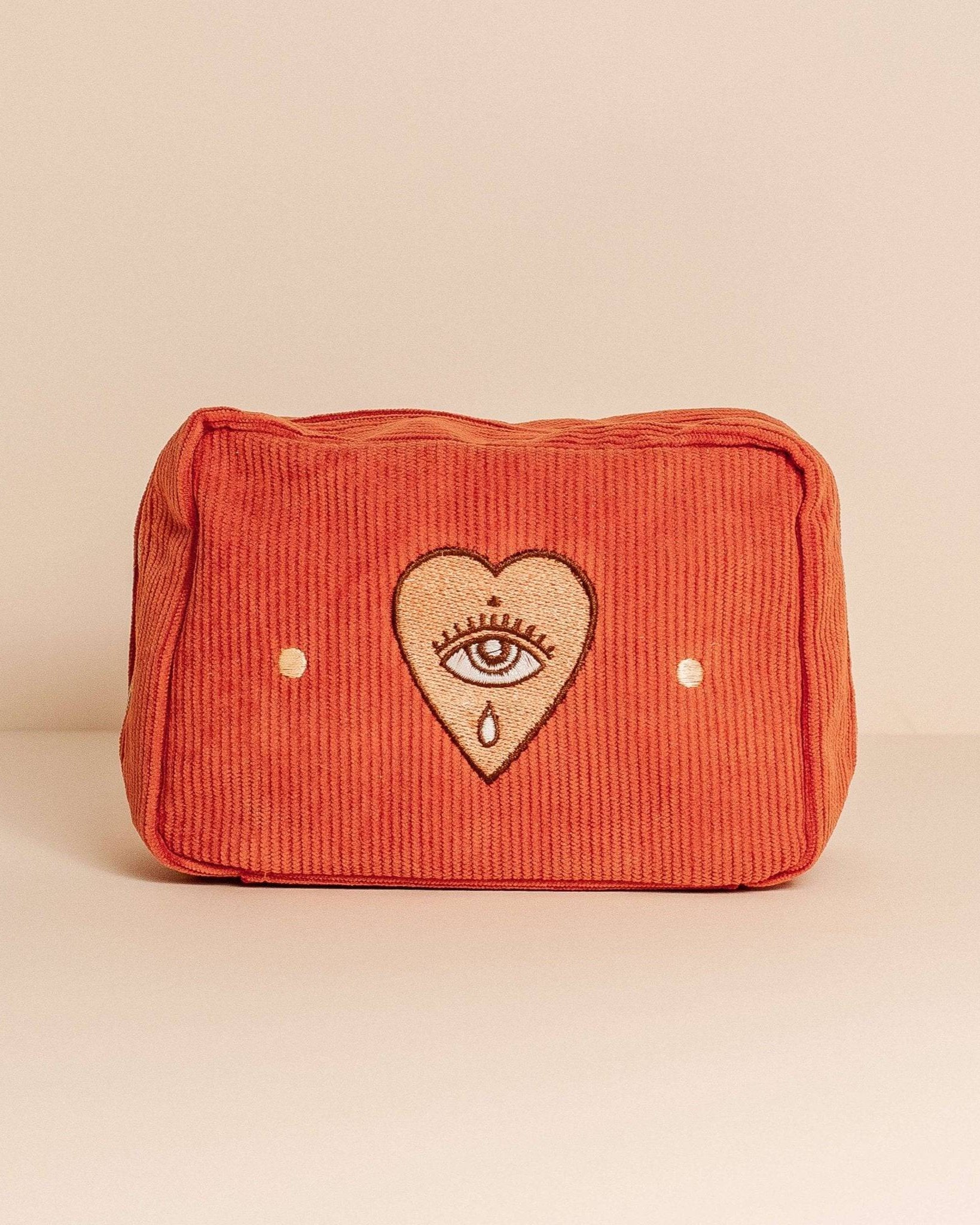 Rust-coloured corduroy makeup bag with a zip closure, fully lined, and five internal pockets. Features a pale orange heart with an eye design.