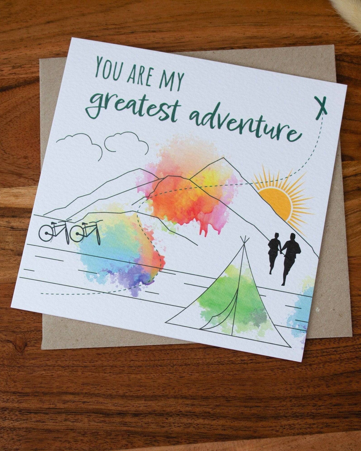 Handmade square greeting card with "You Are My Greatest Adventure" text, featuring a couple running up a mountain in line art with watercolour splashes. Eco-conscious and packaged with a Kraft envelope.