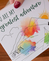 Handmade square greeting card with "You Are My Greatest Adventure" text, featuring a couple running up a mountain in line art with watercolour splashes. Eco-conscious and packaged with a Kraft envelope.