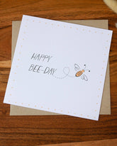 Handmade square birthday card featuring a bumblebee illustration with the words "Happy Bee-Day," printed on premium 100% recycled paper with a biodegradable cello wrap and Kraft envelope.