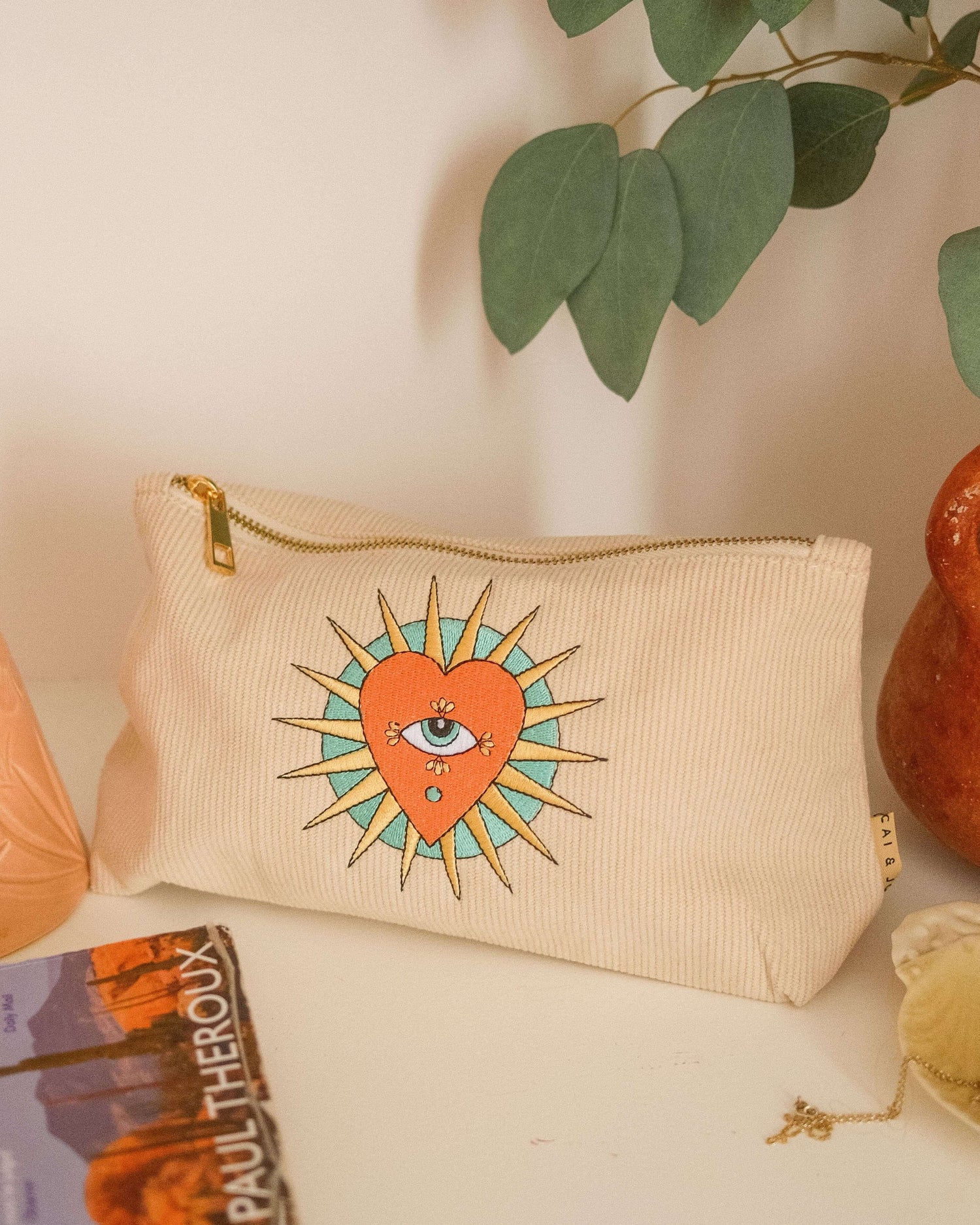 Stone-coloured medium corduroy pouch with a zip closure, featuring embroidered designs of a heart, eye, and sun. Fully lined for storing makeup, toiletries, or travel accessories.