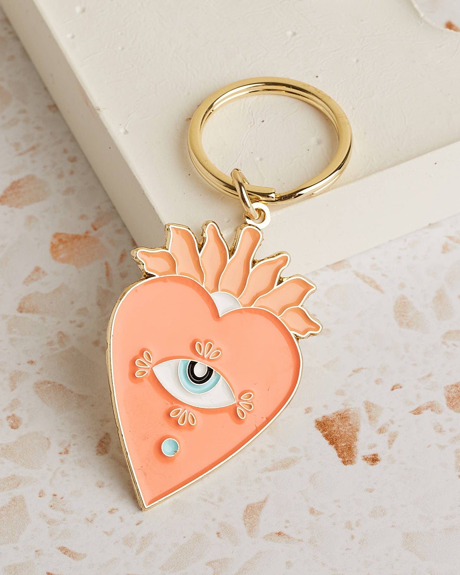 Soft enamel keychain with a pale orange heart design featuring an eye in the centre, accented with gold highlights. Includes a matching gold ring.