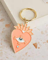 Soft enamel keychain with a pale orange heart design featuring an eye in the centre, accented with gold highlights. Includes a matching gold ring.