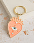 Soft enamel keychain with a pale orange heart design featuring an eye in the centre, accented with gold highlights. Includes a matching gold ring.
