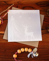 Square handmade Christmas card featuring a minimalist reindeer design in pale blue-grey tones with snow and trees, made with sustainably sourced textured paper and eco-friendly packaging.