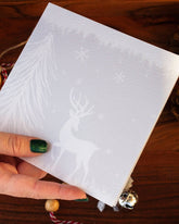 quare handmade Christmas card featuring a minimalist reindeer design in pale blue-grey tones with snow and trees, made with sustainably sourced textured paper and eco-friendly packaging.