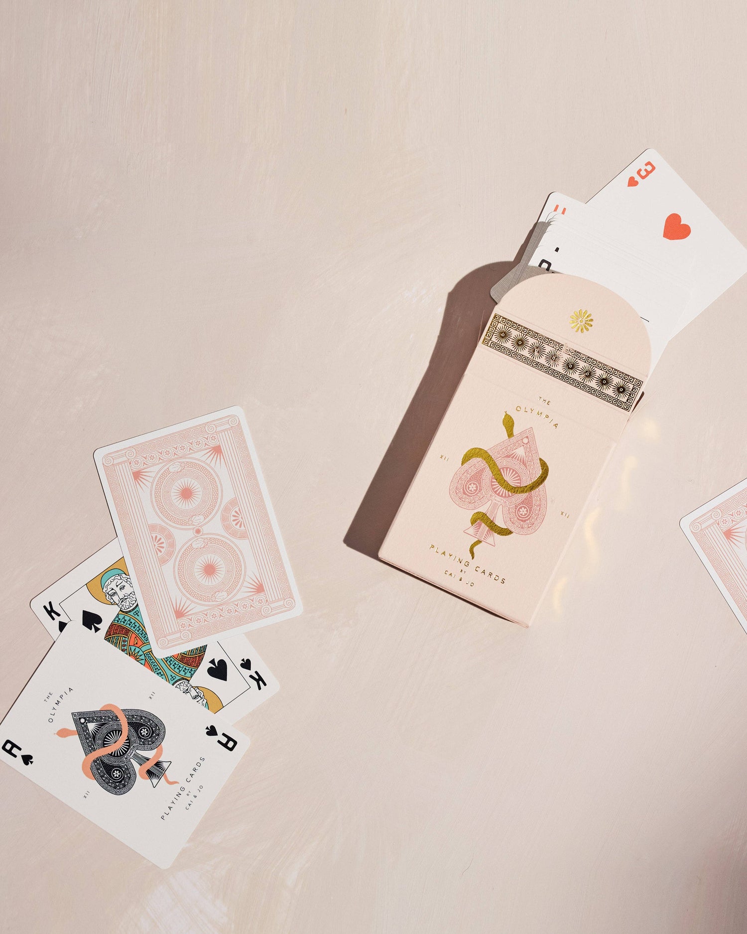 Premium-grade playing cards in pale pink with a gold-foiled tuck box featuring an ace of spades design with a snake wrapped around it. Perfect for games and gifting.