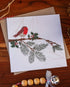 Handmade square Christmas card featuring a robin perched on a holly branch, printed on sustainably sourced textured paper with a biodegradable cello wrap and Kraft envelope.