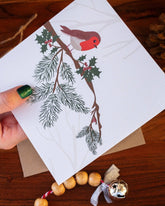 Handmade square Christmas card featuring a robin perched on a holly branch, printed on sustainably sourced textured paper with a biodegradable cello wrap and Kraft envelope.