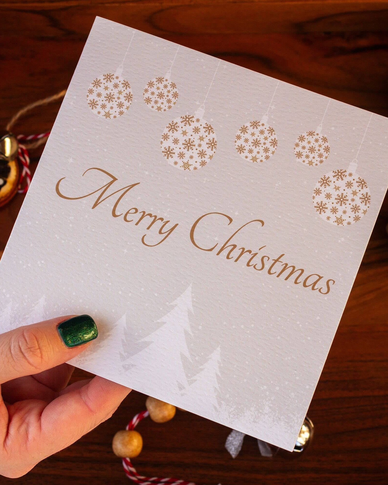 Square Christmas card with white snowy trees, hanging gold and white baubles, and "Merry Christmas" written in gold. Made from recyclable luxury textured paper with eco-friendly packaging.