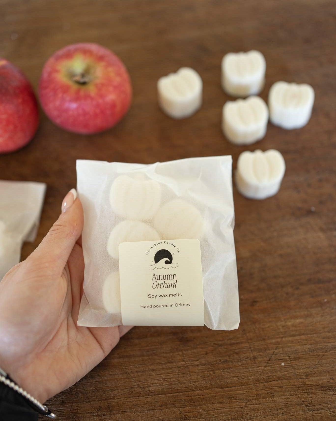 Hand holding a package of soap with apples and soap bars in the background on a wooden surface