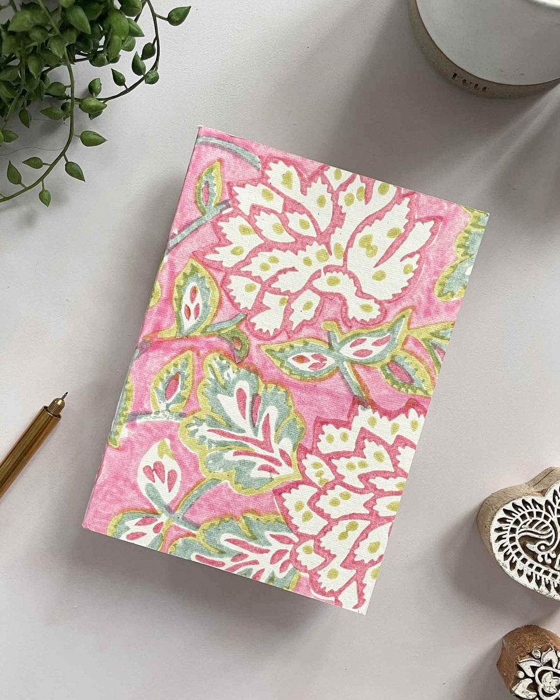 Floral notebook on a white surface with a pen, plant, and heart-shaped stamp.
