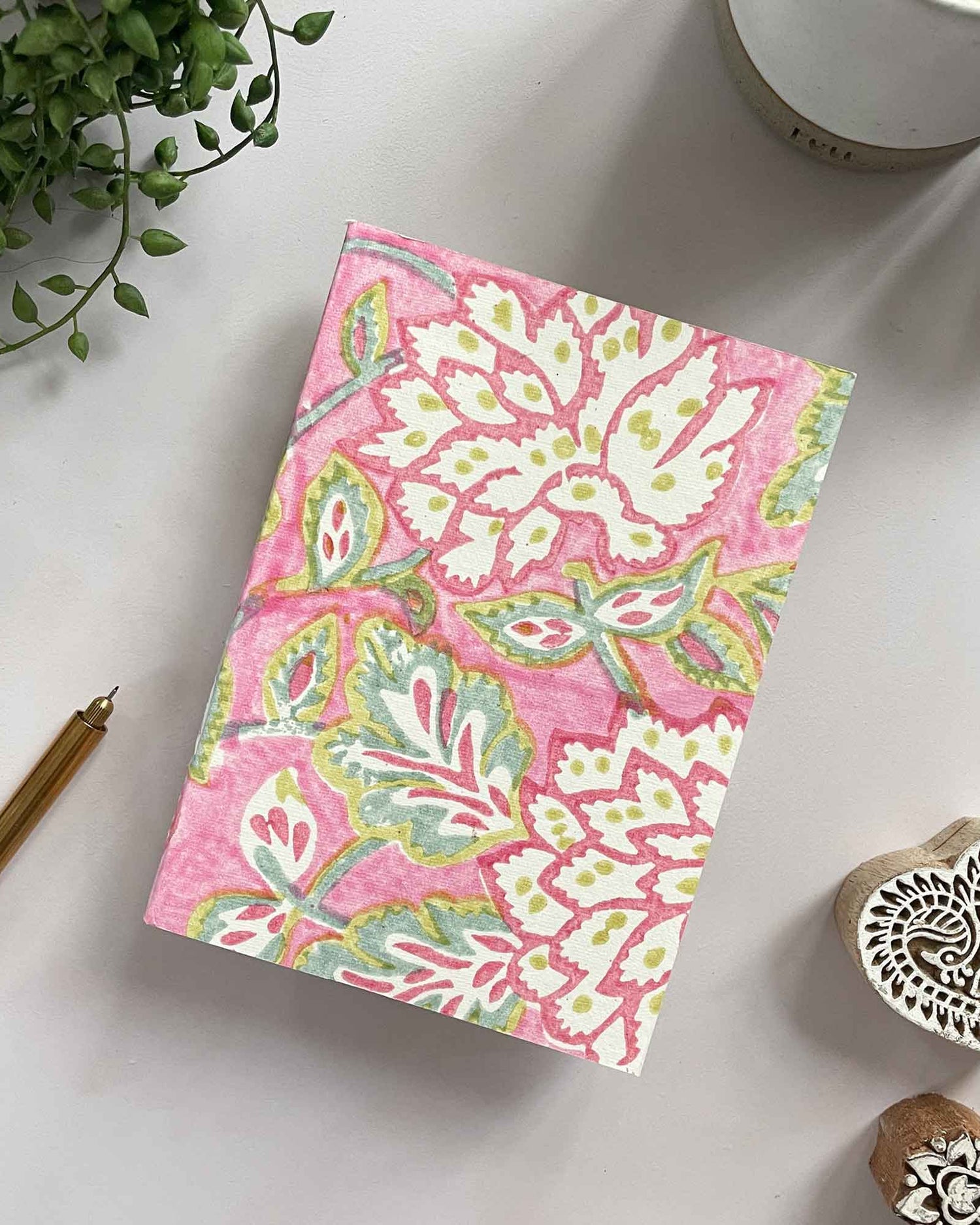 Floral notebook on a white surface with a pen, plant, and heart-shaped stamp.