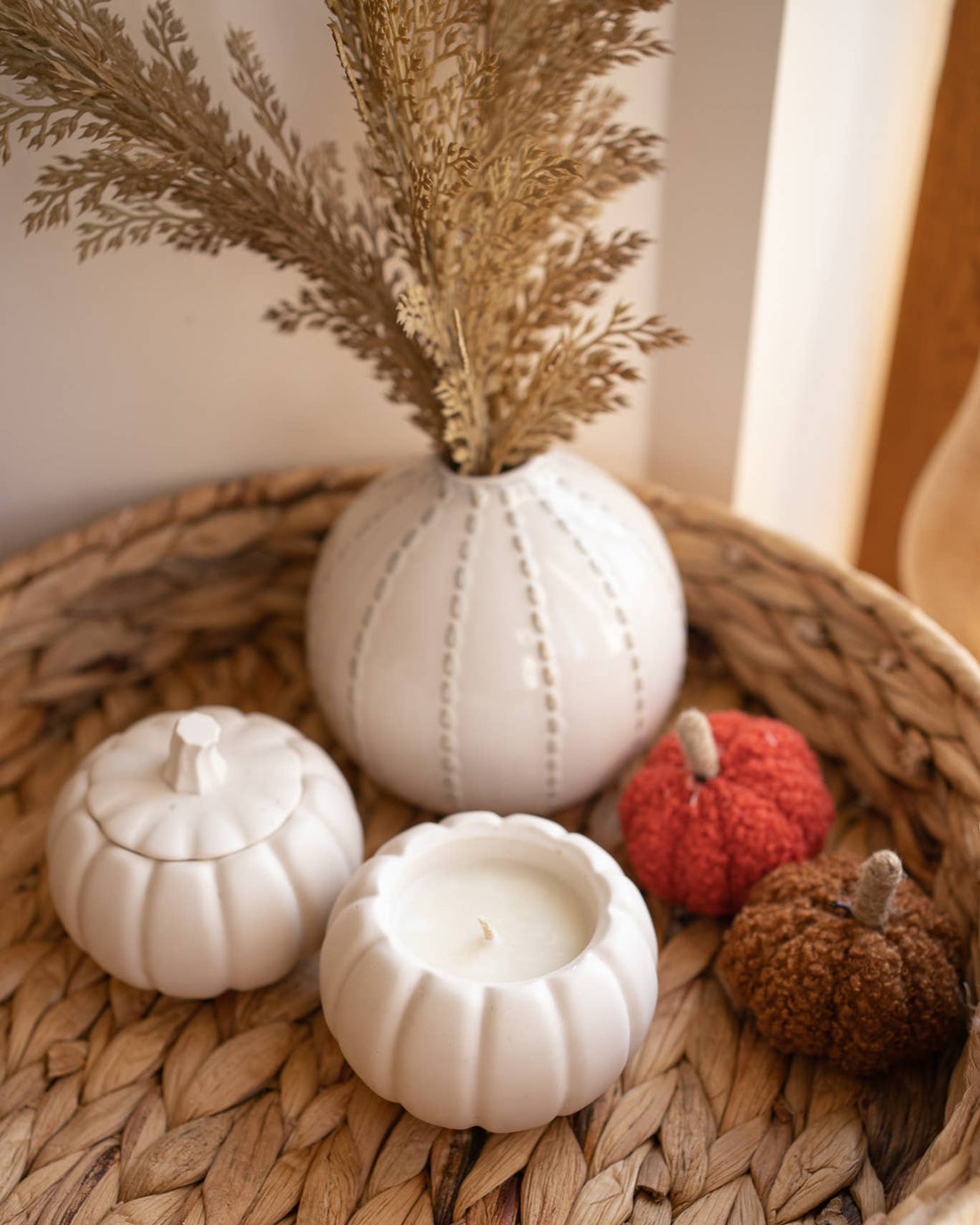 Decorative arrangement with white textured vase, small pumpkins, and dried grasses on a woven surface.