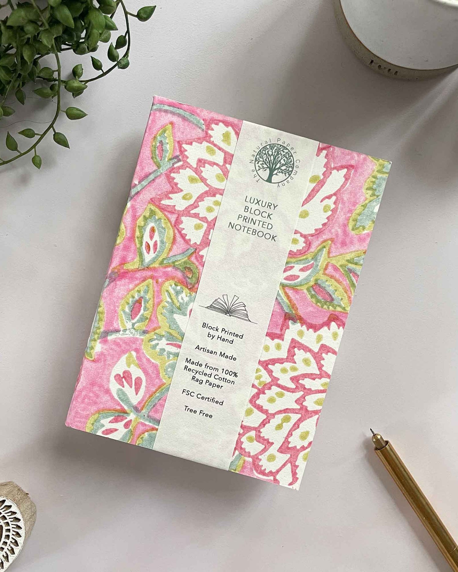 Decorative notebook with pink floral pattern on a white surface with a plant and pen.