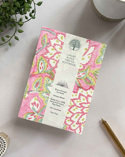 Decorative notebook with pink floral pattern on a white surface with a plant and pen.