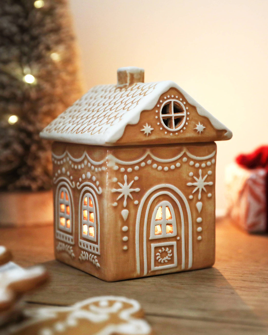 Decorative gingerbread house lantern on a wooden surface with blurred Christmas tree and gift in the background.