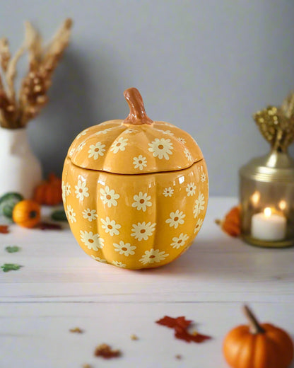 Yellow pumpkin-shaped container with floral design on a wooden surface, next to pink spiral candles.