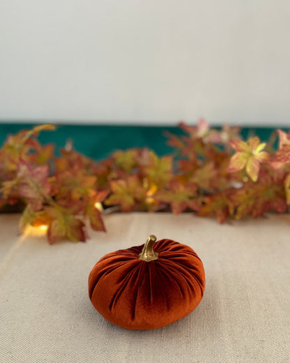 Small orange velvet pumpkin on a beige surface with autumn leaves in the background.