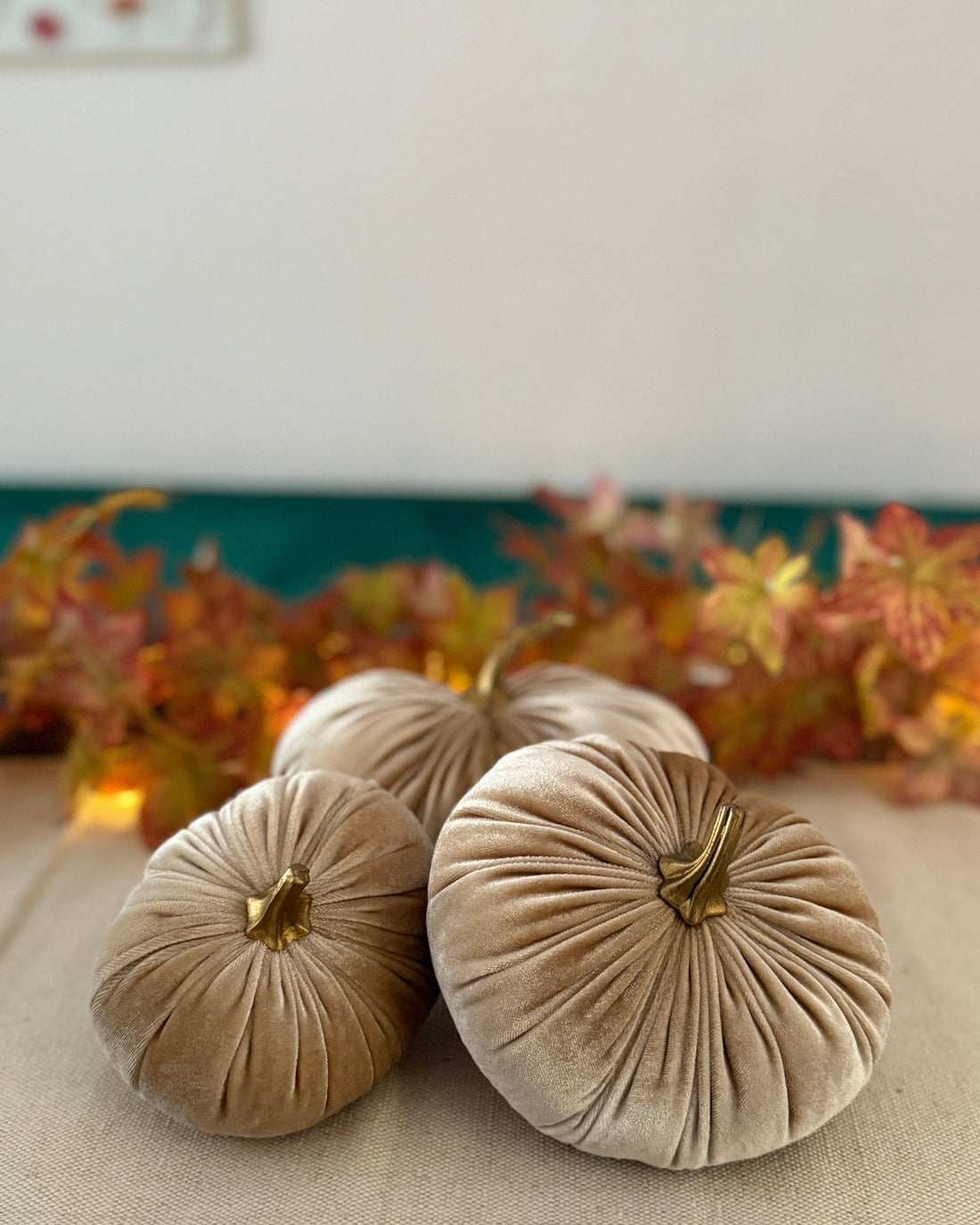 Three beige velvet pumpkins on a table with a blurred autumn leaf background