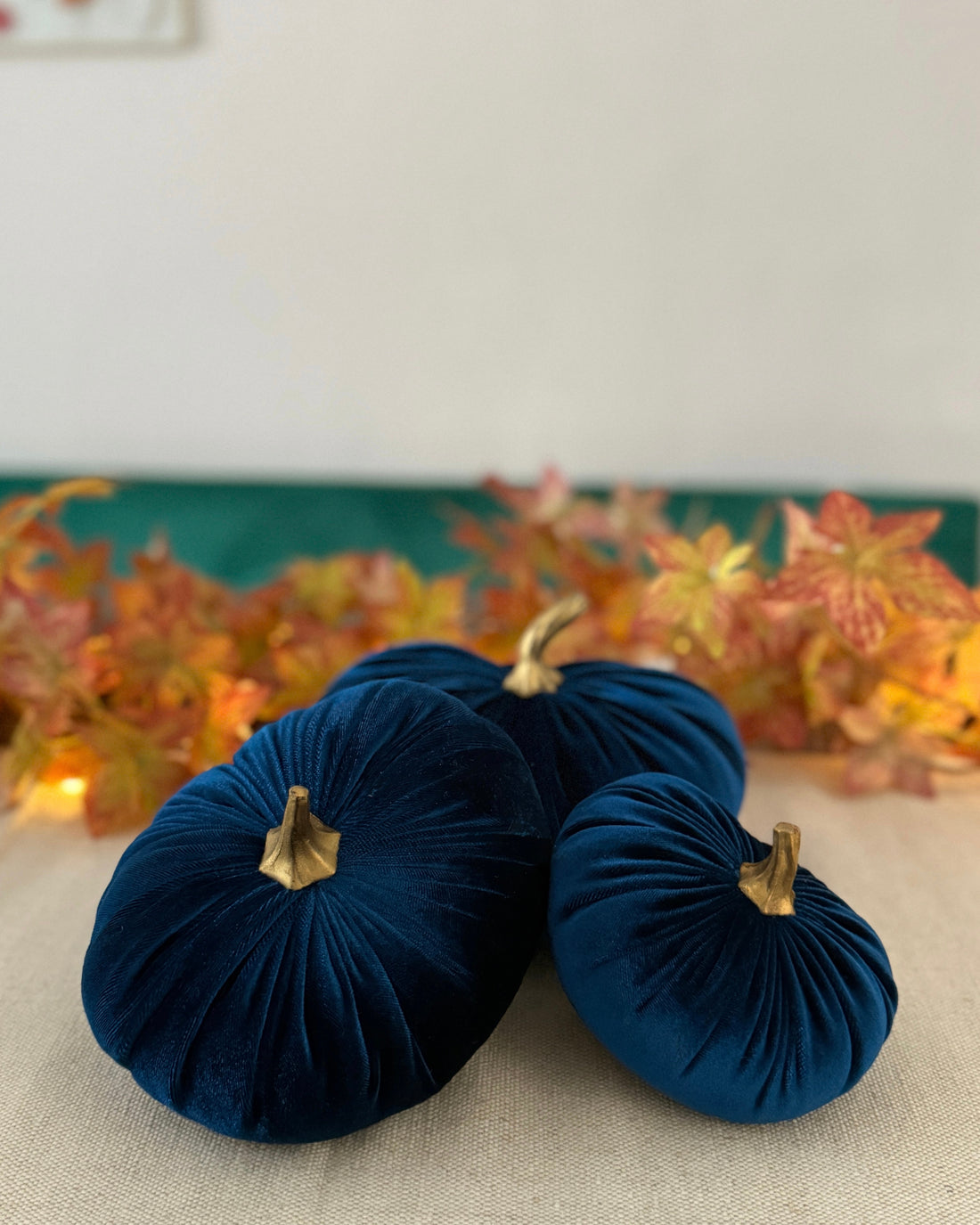 Three navy blue velvet pumpkins with gold stems on a neutral surface with a decorative background.