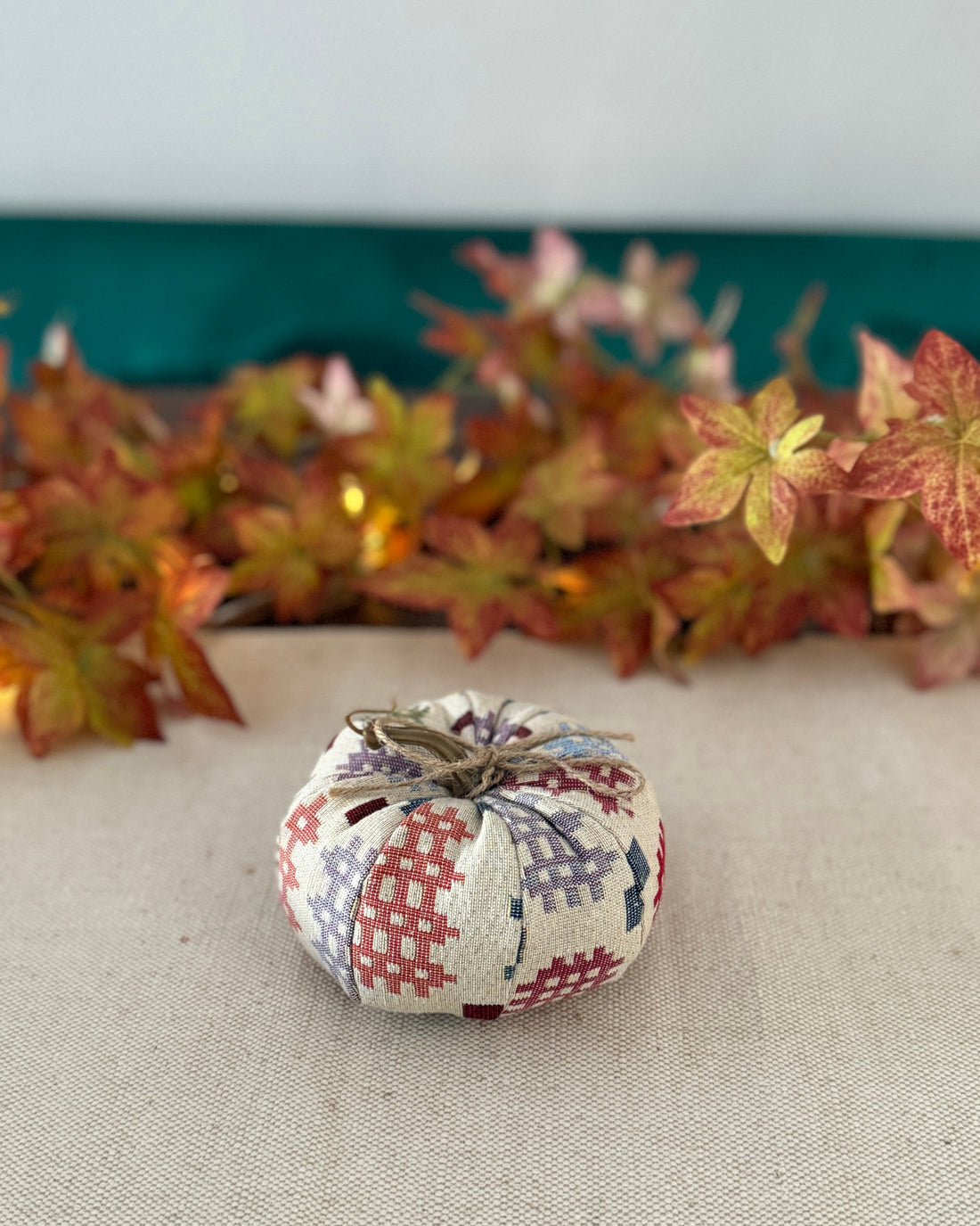 Patchwork pumpkin decoration on a neutral surface with autumn leaves in the background