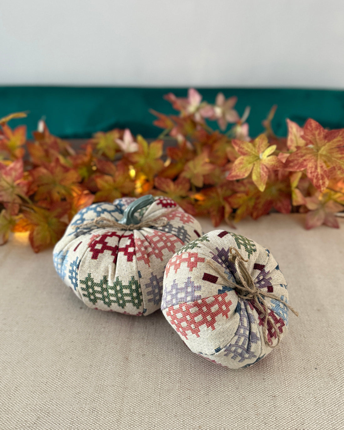 Two patchwork-style fabric pumpkins on a neutral surface with autumn leaves in the background.