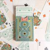 Premium-grade playing cards in sage green with a gold-foiled tuck box featuring an ace of spades design with a snake wrapped around it. Back of box with cards coming out surrounded by cards