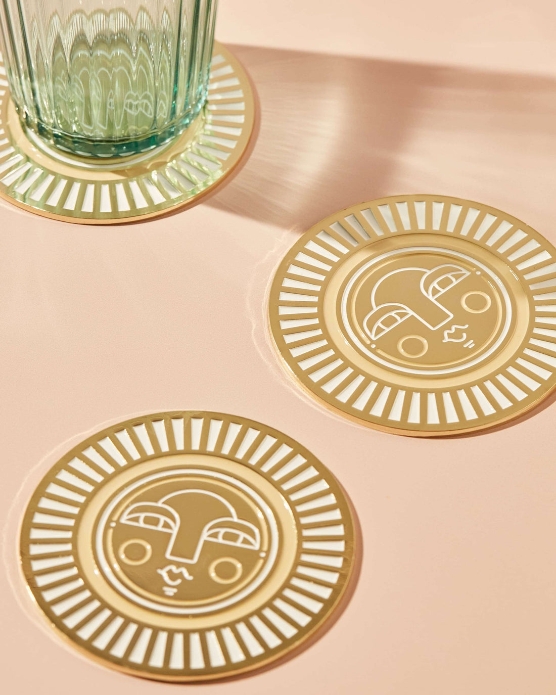 Set of two handmade round warm beige coasters with gold-coloured metal and enamel, showcasing the Sol face design. Ideal for home decor or barware.