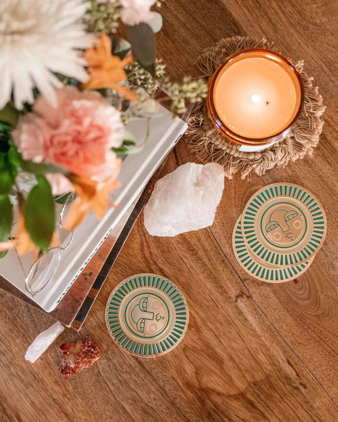handmade teal coasters with gold-coloured metal and enamel, featuring the Sol face design. Coasters placed on a table setting with flowers, candle and books