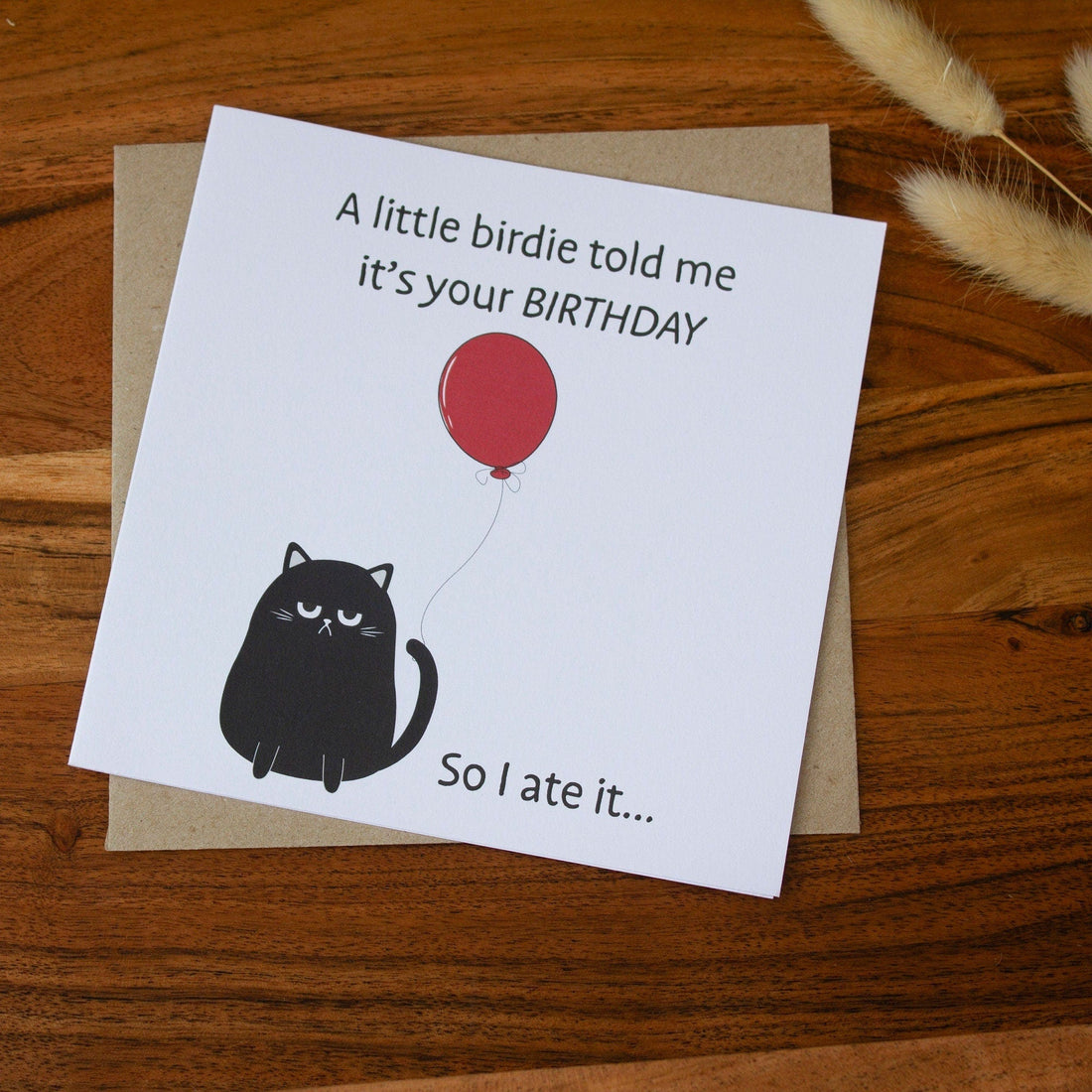 Square birthday card with a humorous black cat illustration holding a red balloon and text: &quot;A little birdie told me it’s your birthday, so I ate it.&quot; Made with sustainable materials.