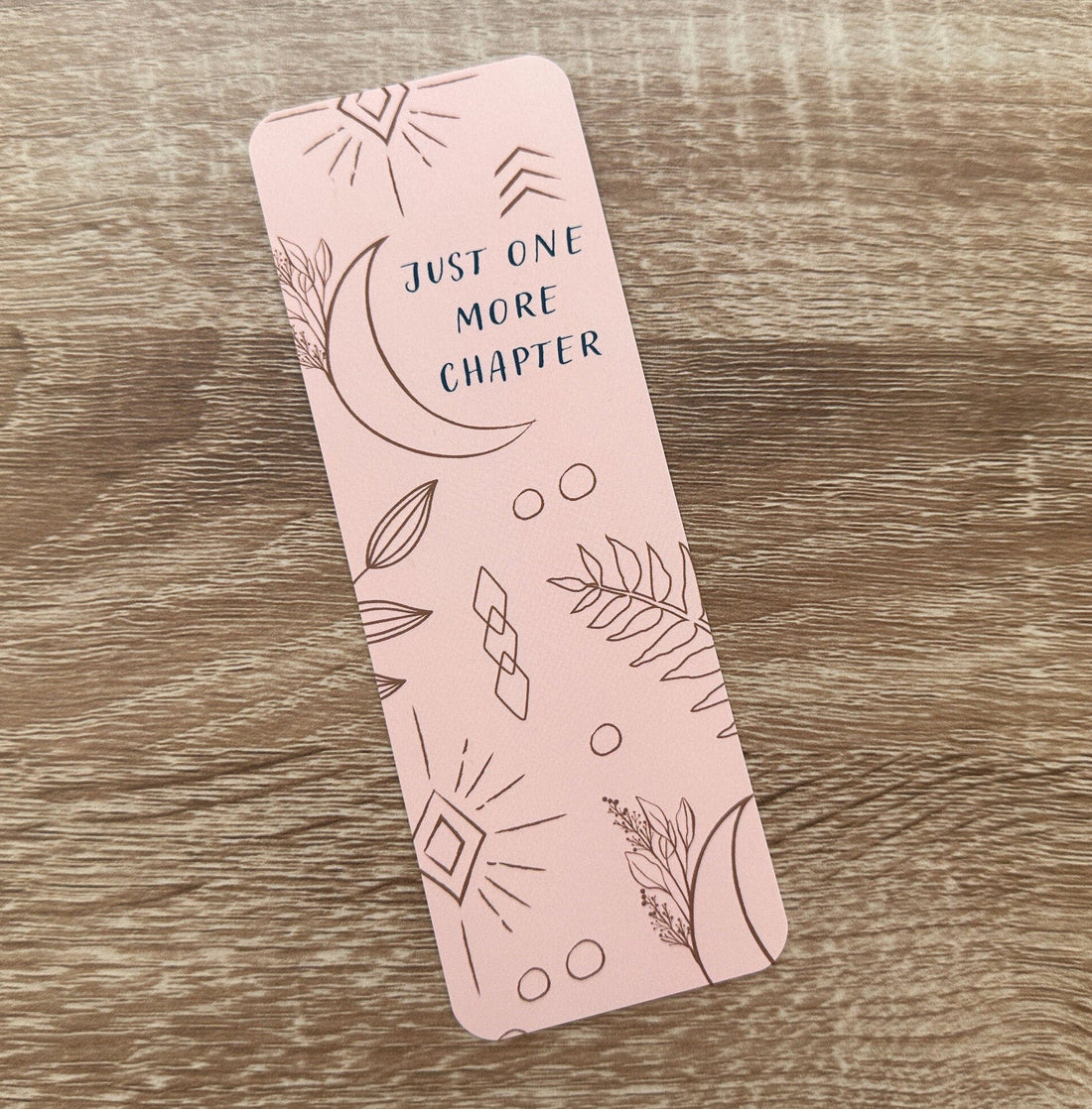 Boho-themed waterproof bookmark with &quot;Just One More Chapter&quot; text, featuring illustrations of moons, leaves, and abstract shapes in pink and brown tones. Made from recycled card with a velvet lamination.