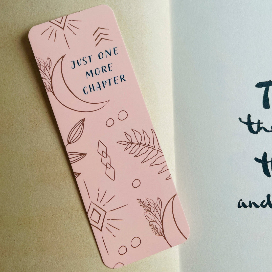 Boho-themed waterproof bookmark with &quot;Just One More Chapter&quot; text, featuring illustrations of moons, leaves, and abstract shapes in pink and brown tones. Made from recycled card with a velvet lamination.