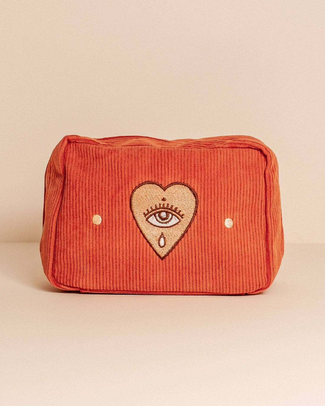 Rust-coloured corduroy makeup bag with a zip closure, fully lined, and five internal pockets. Features a pale orange heart with an eye design.