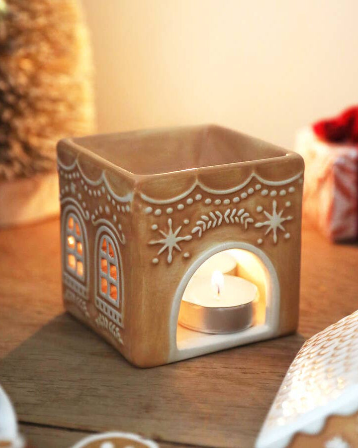 Decorative gingerbread house candle warmer with a lit candle inside on a wooden surface.